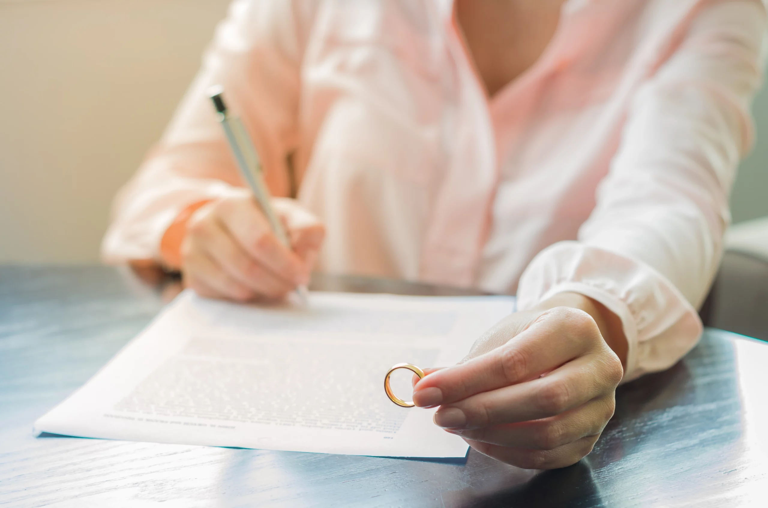Person in a light shirt signing a legal document at a wooden table while holding a gold ring, suggesting a post‑nuptial agreement or important marital decision.