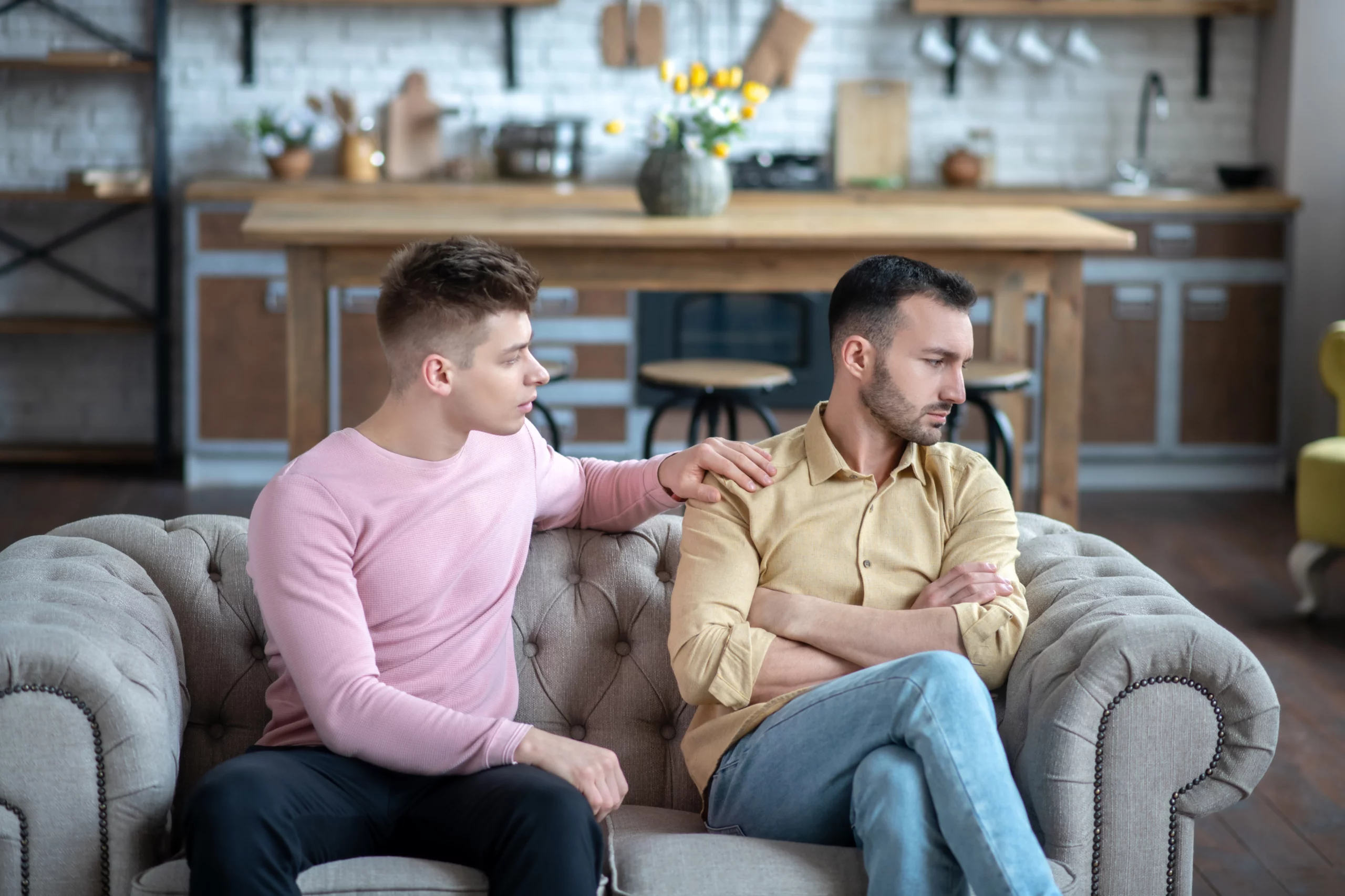Two men on a sofa in a cozy kitchen — one in pink gently consoling the other who seems upset, reflecting emotional support amid a same‑sex divorce.