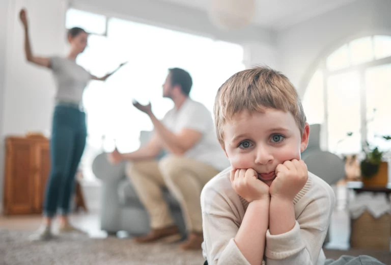 Sad young boy with blue eyes resting his chin on his hands, while a couple argues in the blurred background — illustrating emotional impact of temporary court orders in Florida family law.