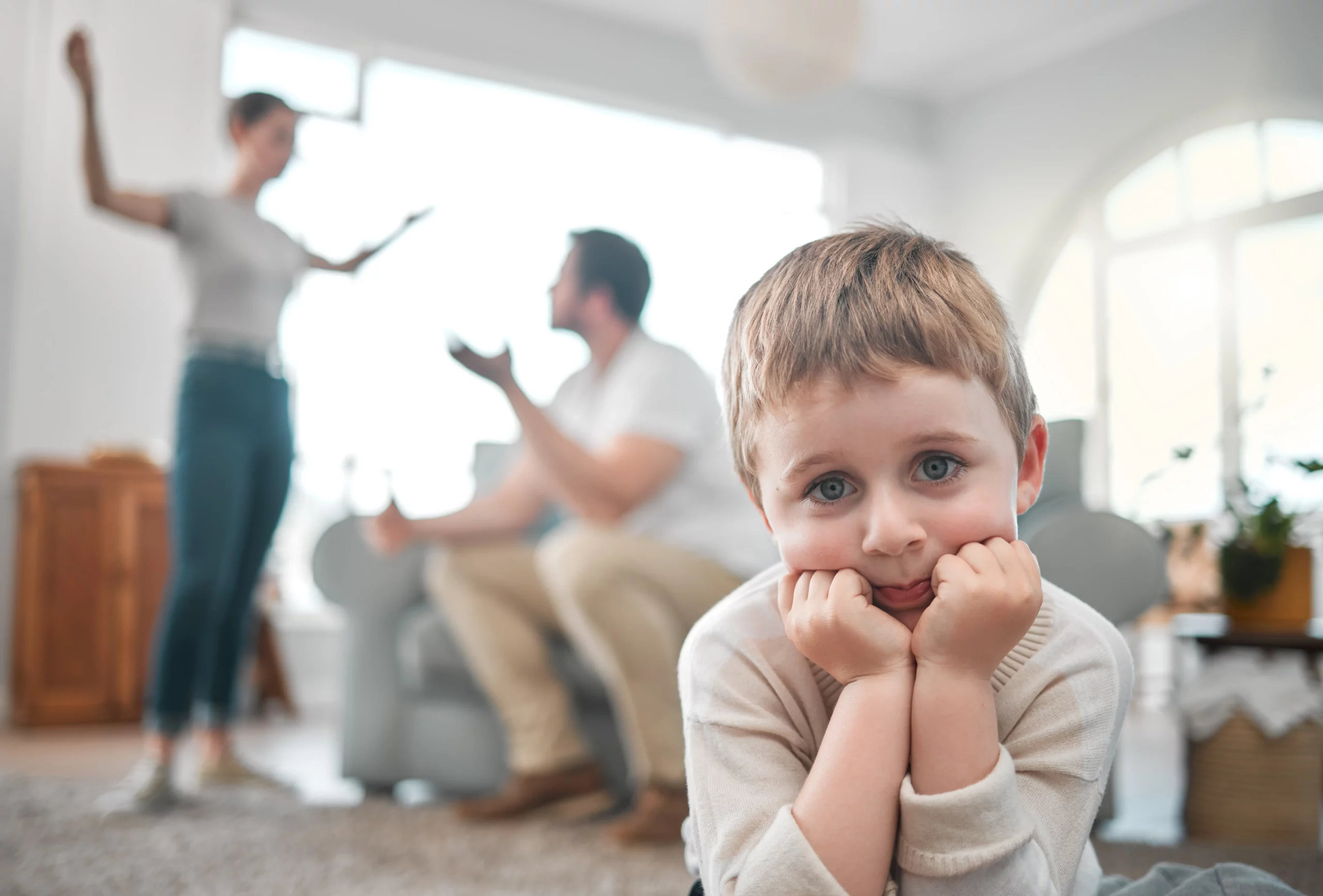 Sad young boy with blue eyes resting his chin on his hands, while a couple argues in the blurred background — illustrating emotional impact of temporary court orders in Florida family law.